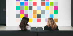 Two women chat in front of a large painting made of colorful squares