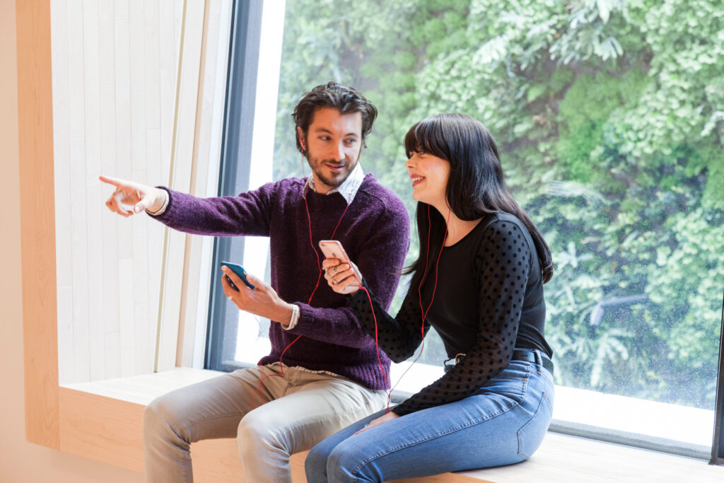 A couple sits on a window bench listening together and pointing