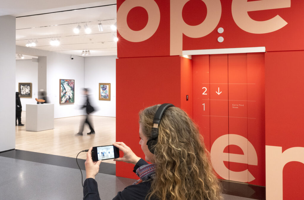 A woman wears headphones attached to a mobile phone that she holds in front of her as she walks the SFMOMA galleries listening to the guided interactive The Telephone Call by Janet Cardiff.