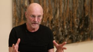 Artist Anselm Kiefer in the SFMOMA galleries with one of his large, dark, textural paintings on the wall behind him. 