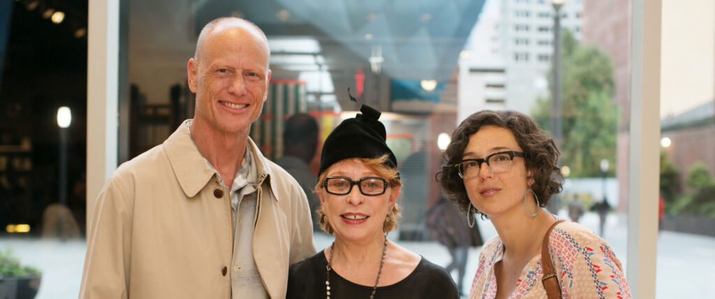 Three people stand together; the woman in the center wears a fancy black hat