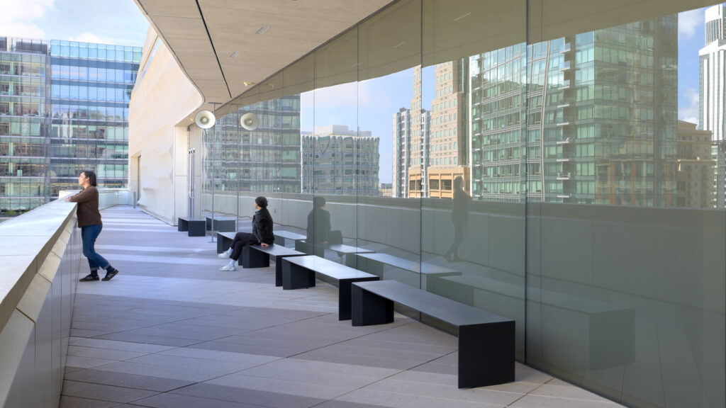 Two people, one sitting and one standing outdoors on the SFMOMA terrace, enjoy the sound installation by artist Susan Philipsz while looking at panoramic views of downtown San Francisco.